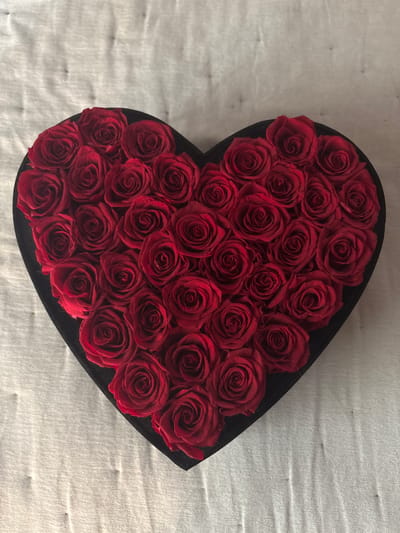 A heart-shaped box filled with red roses sits on a textured, light-colored surface. The box is black and appears to be made of velvet. The roses are tightly packed, with their deep red petals forming a dense arrangement. The background surface has a subtle, woven texture, with some shadowing visible. No people or animals are present in the image. The location is not discernible from the image itself.