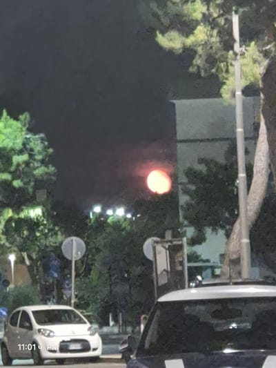 The image depicts a nighttime street scene in Barletta, Italy, with a hazy, red-orange orb visible in the dark sky, likely the moon or a distant light source. In the foreground, a white Citroën C1 car is parked on the right side of the street, partially obscuring a police vehicle. The car has its headlights on, illuminating the street. To the left of the car, a street sign with a white circular marker and a number "1" is visible. Trees and buildings are scattered throughout the background, some with illuminated windows. The overall impression is that of a slightly blurred, atmospheric night scene in an urban setting.