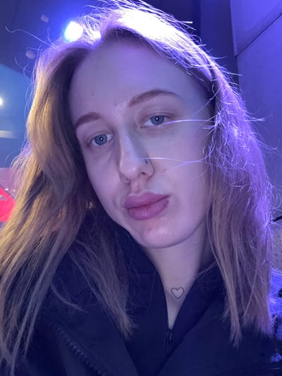 The image shows a fair-skinned woman with blue eyes and light brown hair. She has a nose piercing on the left side. A small heart tattoo is visible on her neck. She is wearing a dark-colored jacket. The lighting is predominantly blue and purple. The location is not identifiable as being in Ol'ginskaya, Russia.