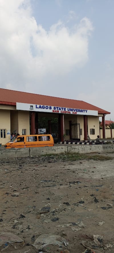 The image displays the main entrance of Lagos State University, Main Campus, Ojo, Nigeria. The foreground consists of an uneven, light brown dirt ground extensively littered with various debris, including plastic bags and discarded materials. A low concrete barrier with black and white striped markings runs horizontally across the mid-ground. Behind this barrier, a bright orange passenger van, containing multiple individuals, is positioned in front of the university entrance. The entrance building features a light-colored facade with multiple columns and a reddish-brown corrugated metal roof. A prominent sign above the entrance reads "LAGOS STATE UNIVERSITY" and "MAIN CAMPUS, OJO" in blue lettering, accompanied by a university crest. Ornate metal gates are visible behind the entrance structure. Several individuals are clustered near the left side of the entrance, and a solitary figure is seated on the ground to the far right, behind the concrete barrier. The sky above is partly cloudy, displaying light blue patches interspersed with white clouds.