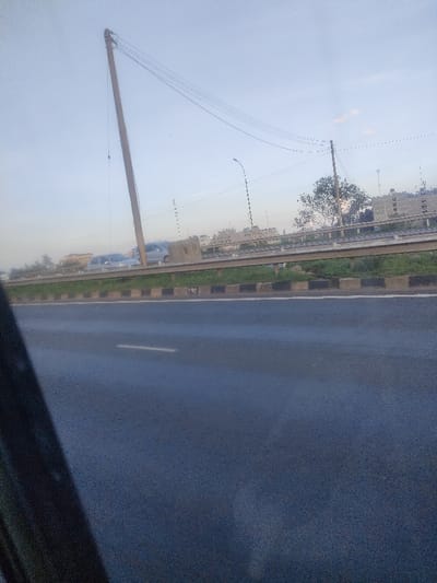 The image displays an outdoor scene in Murera ward, Kenya, featuring a section of roadway and an elevated thoroughfare under a light blue sky. In the immediate foreground, a dark, out-of-focus object partially obscures the lower-left corner of the frame. The road surface, appearing dark grey and possibly damp, features a single white dashed lane marker on the right side. This road is bordered by a concrete curb with alternating black and white painted sections, leading to a grassy median. A metallic guardrail spans horizontally across the midground. Behind the guardrail, an elevated road or structure is visible, carrying two light-colored sedans in motion from left to right. A tall, light-colored utility pole, possibly wooden, stands prominently on the left side of the elevated section, leaning slightly left and supporting multiple overhead power lines that extend towards a shorter utility pole and a green-leafed tree on the right. A street light pole is also visible on the elevated section to the right. In the distant background, a collection of multi-story buildings is observable. No individuals are discernible. The scene is illuminated by natural daylight.