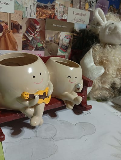 The image shows two decorative ceramic figures resembling eggs with smiling faces and arms and legs, seated on a red bench. The figure on the left holds a miniature yellow guitar. The figure on the right holds a small, shiny object, possibly a ring. Behind the figures, a wall is covered with various collages of photographs, postcards, and text. These include images of a city at night, a retail store interior, stacks of books, and motivational quotes in English and Russian. A fuzzy, white stuffed toy, possibly a rabbit, is partially visible in the background to the right. The figures and bench are placed on a surface that appears to be a desk or table, with a drawing of clouds on paper underneath. The overall scene is a close-up shot, focusing on the ceramic figures and the decorated background. This image does not depict anything related to the city of Kokand, Uzbekistan.