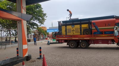 The image shows a red and orange truck with a yellow generator on its back, parked at a petrol station in Sugalimetta, India. The truck has "DHARSAN DRILLING COMPANY" written on its side. A man in an orange shirt is standing on top of the truck, holding pipes. Another man in a blue shirt is standing on the ground near the truck, looking at his phone. In the background, there are trees, a building with a blue roof, and scaffolding. The ground is paved with interlocking bricks. The sky is overcast.