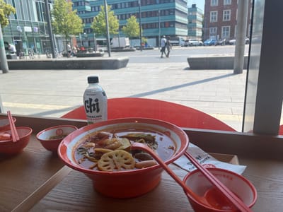A close-up, slightly overhead shot captures a table setting inside a restaurant with a view of a city street in Bottrop, Germany. The foreground is dominated by two bright orange bowls of steaming noodle soup, one larger and in focus, filled with various ingredients including shrimp, lotus root, and possibly meat. Another smaller orange bowl sits to the left, holding chopsticks and a spoon. To the right, a partially visible empty orange bowl contains a spoon. A bottle of "Chi Forest Sparkling White Peach" is positioned behind the bowls. The table surface is made of light-colored wood. In the background, through a large window, a sunlit street scene unfolds, with buildings, trees, a few parked cars, and pedestrians walking. The architectural style of the buildings suggests a modern urban environment. The overall impression is of a casual meal enjoyed indoors with an outward view of city life.