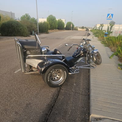 The image shows a black custom trike parked on the side of a road next to a paved walkway. The trike has a large, studded exhaust pipe on the left rear side and a black leather seat and storage compartment. It is positioned with its front wheel on the road and its two rear wheels on the concrete walkway. The background includes a street with lane markings, industrial buildings, trees, and streetlights. A pedestrian crossing sign is visible in the distance on the right. The scene appears to be in an industrial or commercial area of Illescas, Spain, likely during daylight hours with clear skies.