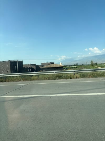The image depicts a roadside scene in Artashat, Armenia. The primary subject is a series of dark, castle-like structures with crenellated battlements and numerous windows, resembling a stylized fortress. These buildings are positioned alongside a paved road, which occupies the right side of the frame and features a white lane marking. A metal guardrail separates the road from a grassy verge dotted with green vegetation.

In the background, a hazy landscape unfolds with rolling hills or mountains under a clear blue sky. Wisps of white clouds are visible, suggesting fair weather. The scene appears to be captured from a moving vehicle, as indicated by the angle and the slight blur in some areas. The time of day is likely daytime, given the bright sunlight casting shadows.

There is no discernible activity or event taking place, with the scene appearing static and focused on the architecture and the surrounding environment. Notable details include the imposing, uniform design of the castle-like structures, the contrast between the dark buildings and the bright sky, and the presence of utility poles and wires in the distance. The overall impression is one of a unique, perhaps tourist-oriented, architectural site located in a semi-rural or roadside setting. No visible text is present in the image.