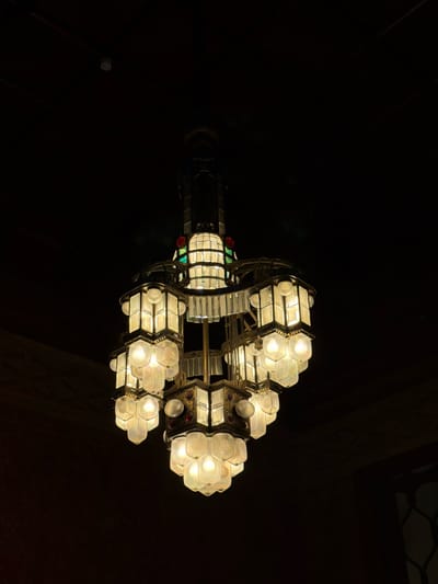 An ornate, multi-tiered chandelier is the central subject of the image, suspended against a dark background. The chandelier is made of brass and features several hexagonal light fixtures with frosted glass panels, illuminated from within, emitting a warm, yellow glow. Some of the fixtures also incorporate faceted clear glass and small, colored glass accents, appearing to be red and green. The chandelier's structure is complex, with vertical elements and cascading light clusters. The background is predominantly dark, suggesting an indoor setting with low ambient light. The upper portion of the image shows a dark ceiling with some faint horizontal lines, possibly beams or architectural features. The lower portion of the image transitions into a dark, textured wall, with a hint of decorative grillwork on the far right. The image appears to be taken from below, looking up at the chandelier. There are no people visible in the image. The overall impression is of a historical or decorative lighting fixture in an dimly lit interior space. The location is identified as the Grand Hotel, San Pellegrino Terme, Italy, known for its Art Nouveau architecture.