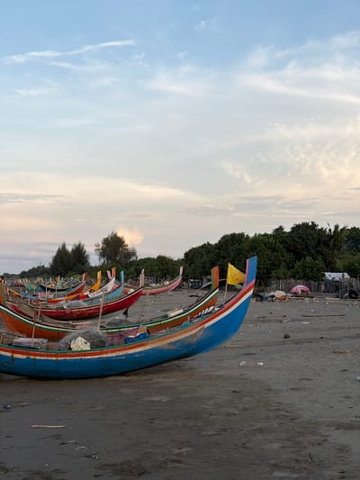 The image depicts a serene beach scene at Pasi Rawa, Indonesia, under a softly clouded sky. Numerous colorful fishing boats are pulled ashore, resting on the dark sand. The boats, featuring vibrant red, blue, orange, and pink hues, are lined up, some with fishing nets and gear visible. In the background, lush green trees and vegetation form a dense line, contrasting with the open sky and the sandy shoreline. Debris, including plastic and tires, is scattered across the beach, hinting at human activity and potential environmental concerns. The overall atmosphere is peaceful, with the gentle light suggesting either early morning or late afternoon.