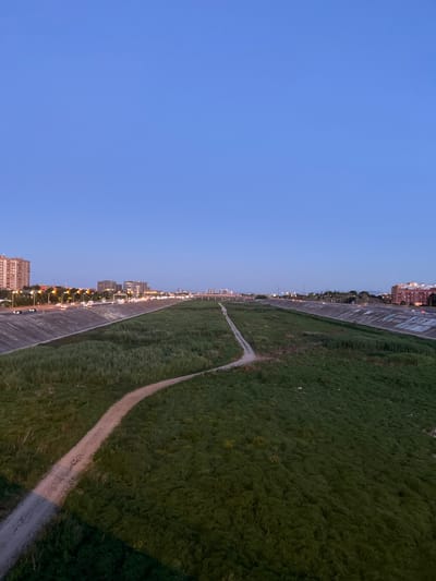 The image depicts a wide, drained riverbed filled with lush green grass and a dirt path winding through it. On the left bank, a busy road with traffic lights and cars is visible, bordered by tall buildings. The right bank is also lined with concrete structures and some graffiti. In the distance, a city skyline with more buildings and bridges can be seen under a clear blue sky. This scene is located in Xirivella, Spain, along the Turia riverbed.
