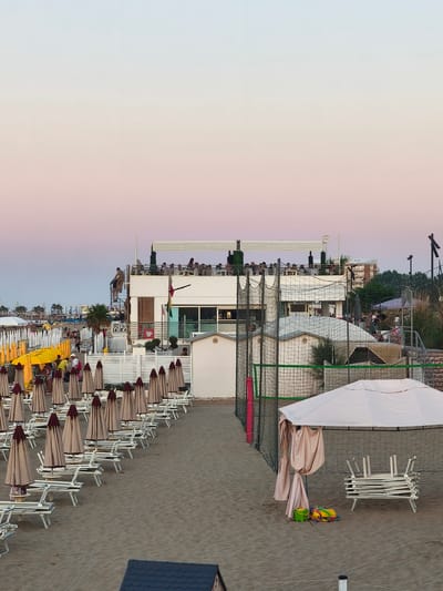 The image depicts a beach scene in San Salvo, Italy, during dusk. The sky exhibits a gradient of colors, transitioning from light blue at the top to soft pink and lilac towards the horizon. The beach is lined with numerous rows of closed beach umbrellas and loungers. In the middle ground, a multi-story building with a rooftop area is visible, with people gathered on the rooftop and on a balcony. A volleyball net setup is present on the sand, with fencing and netting enclosing the court. A large white canopy tent with draped fabric stands on the right side of the image, with a stack of folded white loungers beneath it. In the foreground, a dark blue tiled roof of a small structure is visible. The sand is the primary ground cover. The overall atmosphere suggests a beach resort winding down for the evening.