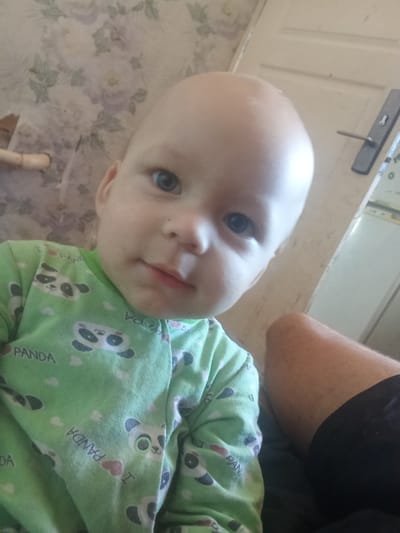 The image is a medium shot, taken indoors, with a shallow depth of field. The subject is a baby, approximately 1-2 years old, bald with light skin and blue eyes. The baby is wearing a light green pajama set with a panda print and the text "PANDA" repeated. The baby is looking directly at the camera with a neutral expression. To the right of the baby, a person's leg is visible, with a tanned thigh and black shorts. In the background, a floral patterned wallpaper is visible on the left, and a white door with a dark handle and a white appliance on the right. The lighting is soft, coming from the front. There is no information in the image to determine the specific location within the city of Дружба, Russia.