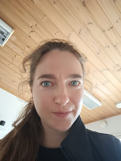 An overhead, close-up, eye-level selfie of a woman with light brown hair and blue eyes is captured in a room with a wooden slatted ceiling. The woman is facing forward, looking directly at the camera with a neutral expression. She is wearing a black jacket and a thin gold necklace. In the background, two ceiling lights are visible, one on the left side of the frame and another on the right. The room appears to be an indoor space, possibly a home or office. The location of Tuusniemi, Finland is not visible or indicated in the image.