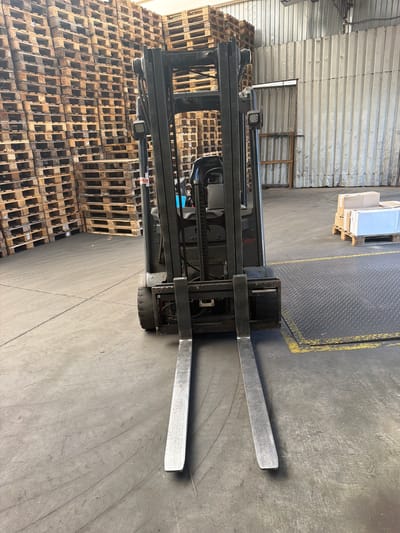 A forklift is parked in a warehouse, facing forward, with its forks lowered to the ground. The forklift is dark gray and appears to be electric. In the background, there are stacks of wooden pallets, a corrugated metal wall, and a closed door. To the right, there is a pallet with boxes. The floor is concrete, with some yellow markings. The image does not provide any information about the location in the city of Olomouc, Czechia.
