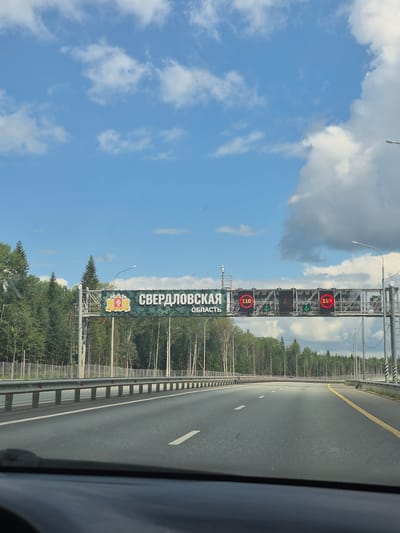 The image shows a view from a vehicle traveling on a highway. A large overhead sign is visible, displaying the words "СВЕРДЛОВСКАЯ ОБЛАСТЬ" (Sverdlovsk Oblast) in Russian, along with the coat of arms of Sverdlovsk Oblast. The sign also features digital displays showing "110" in red circles, indicating a speed limit. Green arrows are also displayed below the speed limit signs, pointing downwards, suggesting lanes ahead. The highway is lined with metal guardrails and trees on both sides. The sky is blue with scattered white clouds. The location is identified as the city of Verkh-Shurtan, Russia.