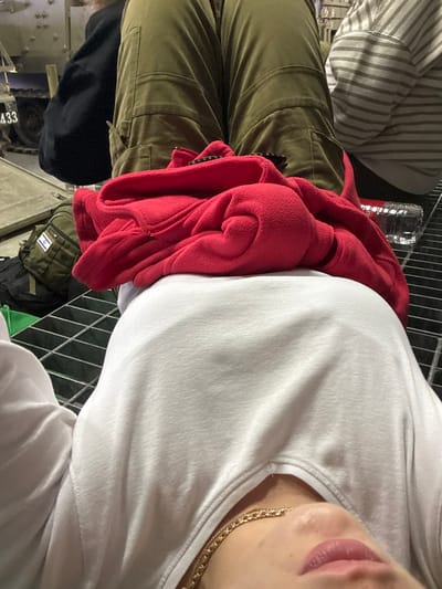 The image depicts a low-angle view of a scene in Rosh Haayin, Israel. In the immediate foreground, the lower face, neck, and upper torso of an individual are visible, wearing a white long-sleeved top and a gold-colored chain necklace. A red fleece jacket is folded and rests on the individual's midsection. Beyond this, a metal grating surface forms the ground. On this grating, a clear plastic water bottle is present. Further back, two other individuals are partially visible: one wearing olive green cargo-style pants, indicative of a military uniform, and another wearing horizontally striped pants. A tactical bag, olive green with an Israeli flag patch, rests on the grating near the individual in military pants. In the background, a light-colored military vehicle, possibly an armored personnel carrier or tank, is partially visible, bearing the numbers "433" on its side. The setting suggests a military or operational environment.