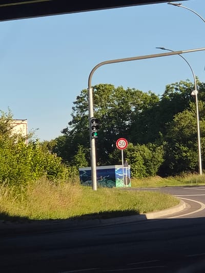 The image shows a traffic light with a green light illuminated, indicating that traffic can proceed. To the right of the traffic light, a circular speed limit sign displays "50" within a red border. The traffic light and speed limit sign are positioned near a road intersection, with trees and vegetation forming the background. A colorful mural is visible on a structure behind the speed limit sign. The scene is set outdoors under a clear blue sky, suggesting daytime. The location is identified as Dudelange, Luxembourg.
