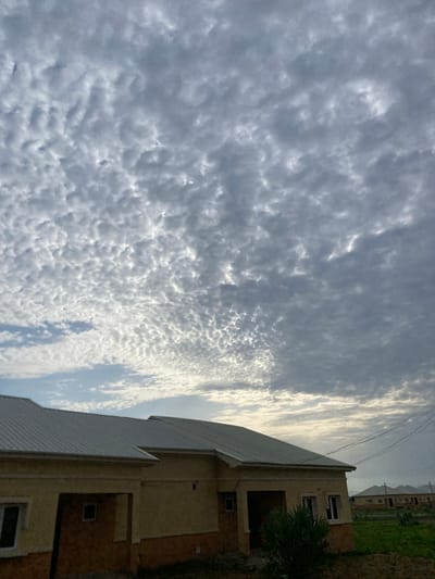 A low-angle, full shot captures a residential area under a dramatic, overcast sky, characteristic of Bulamare, Nigeria. The foreground is dominated by the light brown, stucco facade of two attached houses, featuring silver, corrugated metal roofs. Multiple small, dark-paned windows punctuate the walls, with a darker recessed entryway visible. A dense, dark green shrub partially obscures the right side of the closer house. In the background, a row of similar houses stretches into the distance, their roofs also gleaming silver. Utility wires are visible in the upper right corner, stretching across the frame. The sky is filled with a textured pattern of altocumulus clouds, varying in shades of gray and white, with patches of brighter sky peeking through. The overall impression is of a developing suburban or semi-rural landscape on a cloudy day.