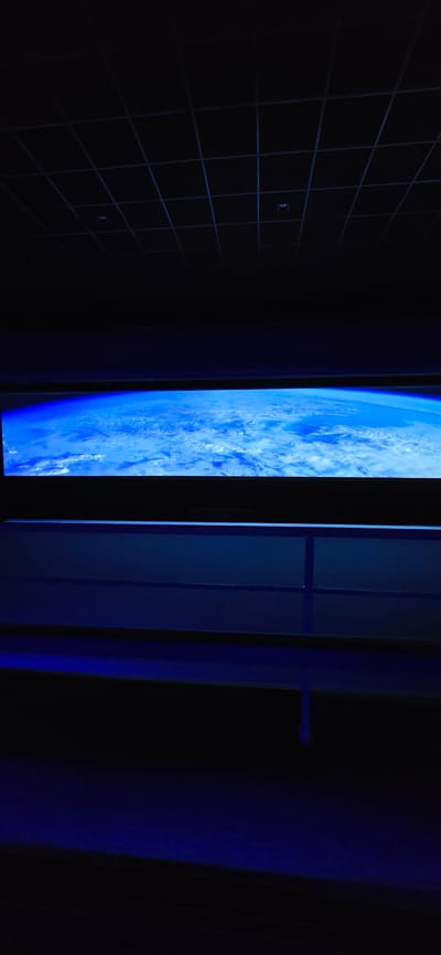 The image appears to be an indoor shot, predominantly dark with blue lighting. On the left, a wall with a grid pattern is visible. In the center, there is a screen displaying an image of Earth from space, with prominent blues and whites representing oceans and clouds. To the right of the screen, vertical panels are visible, seemingly part of a doorway or architectural structure. A handle is visible. Due to the lack of context, the location in Canela, Brazil cannot be confirmed.