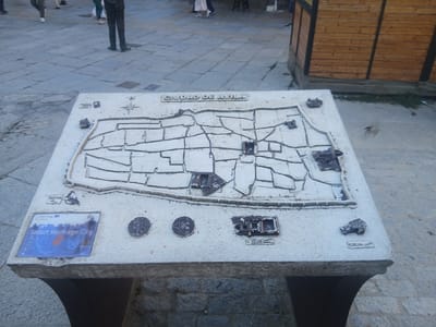 This media file shows a 3D model map of the city of Ávila, Spain, displayed on a stone pedestal outdoors. The map depicts the city's layout with its walls and streets in relief. Several small, raised metal models representing buildings are placed on the map, and a compass rose is visible towards the top left.
On the front left corner of the pedestal, there is a blue and white plaque with text that reads "Smart Heritage City" and "EU" with the European Union flag. Below this, there are two circular metal markers, one with an icon of a sound wave and the text "N", and another with an icon of a building and the text "N". Further to the right on the pedestal, there are other small metal structures and a sign with some text, possibly indicating buildings or points of interest. A bird is captured in mid-flight to the right of the map.
In the background, the ground is paved with cobblestones. Several people are walking in the background, suggesting a public space like a plaza or street. A wooden structure with dark framing is visible on the right side of the frame, partially obscuring the background. The lighting suggests it is daytime, possibly late afternoon given the shadows. The overall scene appears to be a tourist information point or an outdoor exhibit within the city.
