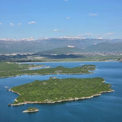 Aerial view of Lake Skadar in Ponikvica, Montenegro. The lake is blue, and there are several green islands. A mountain range is visible in the background. The sky is blue with some clouds. A narrow strip of land and a small town or village are situated between the mountains and the lake.