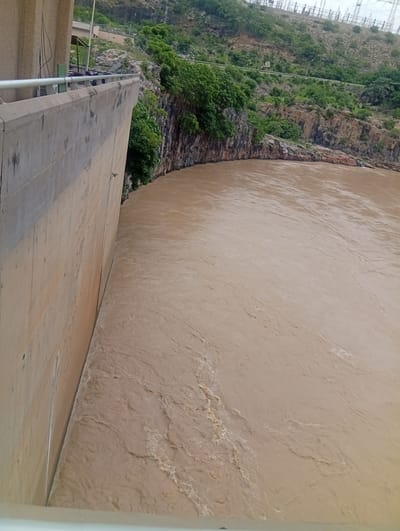 An elevated, eye-level, full shot captures a dam wall made of concrete, with a wide body of murky, brown water flowing beside it. The dam wall is on the left of the frame, angled inward toward the top of the image. The water occupies the right two-thirds of the image, with ripples and currents visible across its surface. The background features a rocky hillside covered in green vegetation, with the top of the hillside showing electrical towers. The scene is located at the Shiroro Dam in Shiroro, Nigeria.
