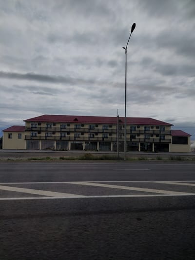 A long, three-story building with a red roof and cream-colored walls stands beside a road under a cloudy sky. The building appears to be a hotel or motel, with numerous windows and balconies. A tall street lamp is visible in front of the building, and a road with white markings stretches across the foreground. The scene is located in Usharal, Kazakhstan.