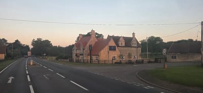 The image captures a scene at dusk on a rural road in Thorpe Waterville, United Kingdom. In the foreground, the asphalt road curves to the right, with white lane markings and an arrow indicating the direction of travel. A temporary traffic cone is visible on a central island in the road.
To the left, a building constructed of stone with a red tiled roof is partially visible. Further down the road, a truck with a red and white trailer and a dark-colored car are traveling away from the camera.
The main subject of the image is a large, traditional stone building, likely a pub or inn, identified by a sign that reads "THE FOX INN" with a graphic of a fox. This building has a red tiled roof, multiple chimneys, and numerous windows. It is surrounded by a dark wooden fence. Adjacent to this building, and also visible in the image, is a smaller, dark-roofed building with a stone wall. A circular speed limit sign indicating "30" is posted on a pole to the right of the inn.
The background consists of a dense line of trees and foliage. The sky is a pale orange and grey, indicative of twilight. Power lines are visible stretching across the upper part of the frame. The overall environment appears to be a tranquil countryside setting.