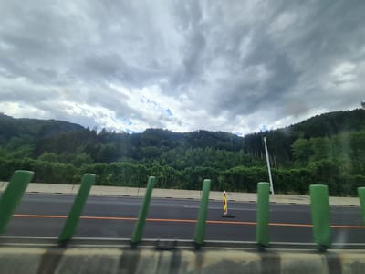 The image shows a view from a vehicle traveling on a highway, likely in the vicinity of Leoben, Austria. The sky is overcast with dramatic, dark clouds. In the foreground, there are several green protective barriers along the side of the road. The highway itself is asphalt with an orange line marking a lane. Beyond the highway, a dense forest covers rolling hills that recede into the distance. A white pole, possibly a lamppost or communication tower, stands on the right side of the frame, partially obscured by trees. A construction cone with red and yellow stripes is visible on the road surface between the green barriers. The overall impression is a cloudy, rural highway scene.