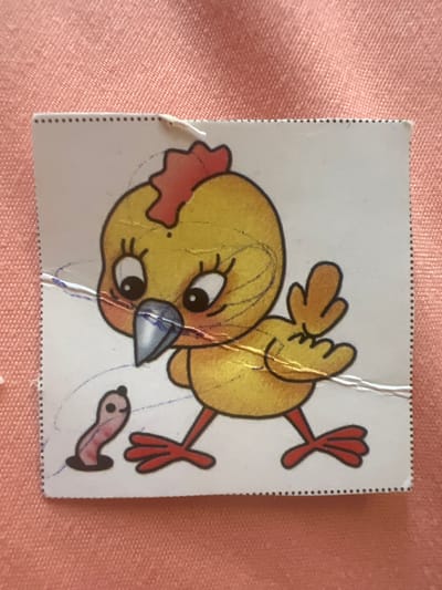 The image is a close-up, high-angle shot of a small sticker featuring a cartoon chick and a worm on a pink, textured surface. The sticker is square with a perforated edge along the top and right sides. The chick is yellow with a red crest, large black eyes with eyelashes, and a blue and gray beak. It has orange feet. A small, pink worm with a black head is emerging from a dark brown hole on the left side of the sticker. The sticker appears to be slightly damaged, with some creases and what looks like faint blue scribbles on it. The pink surface underneath is consistent with fabric. There is no information in the image to suggest any connection to the city of Pskov, Russia.