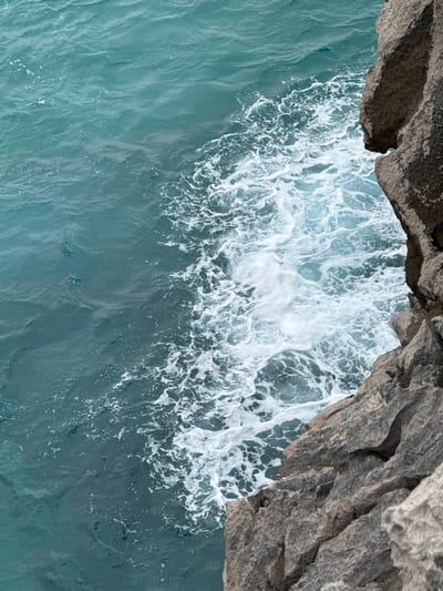 The image depicts a close-up, high-angle view of the turbulent turquoise sea meeting a rugged, grey rocky cliff face. The water is a deep, vibrant blue-green, with frothy white waves crashing and swirling against the rocks. The cliff is textured and weathered, showing cracks and fissures. The scene appears to be a natural coastal environment, likely in Llanes, Spain, known for its dramatic coastline and picturesque beaches. There are no people or man-made objects visible in this frame.