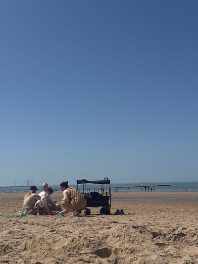 The image shows a beach scene, likely in Knokke-Heist, Belgium, given the distinctive pier-like structures visible in the background. Several people, including adults and children, are gathered on the sand. They appear to be engaged in activities such as playing or building in the sand. A beach cart or wagon is parked nearby, holding items. The weather appears sunny with a clear blue sky. The beach stretches out to the sea, with some people visible in the distance.