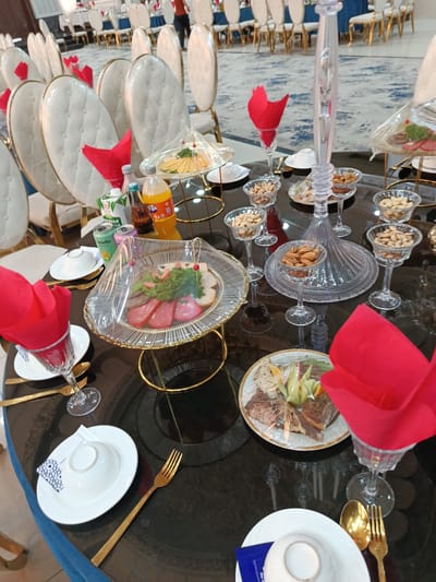 The image depicts a banquet table set for an event, likely a wedding or a formal gathering, in Shahrisabz City, Uzbekistan. The table is covered with a dark, reflective surface, and numerous white chairs with gold trim surround it, suggesting a grand setting. The event appears to be in progress, with place settings for multiple guests, each featuring white plates, gold cutlery, and a red napkin artfully folded in a glass.

Several food items are visible, including platters of sliced meats and cheeses, as well as small bowls filled with nuts such as almonds and pistachios. Beverages, including what appears to be a bottle of orange soda and cans of soft drinks, are also present on the table. A decorative glass centerpiece stands prominently in the middle of the table. The lighting suggests it is daytime, possibly late afternoon or early evening. The overall atmosphere conveyed is one of elegance and celebration. While no people are clearly in focus, a blurred figure can be seen in the background, hinting at the presence of guests and attendees. There is no discernible text in the image.
