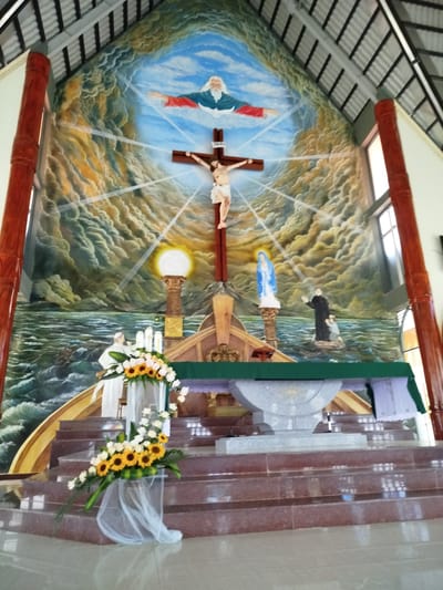 The image depicts the interior of a church in K'Rèn, Vietnam, featuring a large mural and altar. The mural, painted on the back wall, is a religious scene with a depiction of God at the top, Jesus Christ on the cross in the center, and other figures such as Mary and a priest with a child in the lower portion, all set against a backdrop of stormy clouds and water. At the front of the church, the altar is visible, adorned with floral arrangements of sunflowers and white roses, and illuminated by candles. The altar is positioned on a multi-tiered staircase. The architecture of the church, with its pitched roof and wooden pillars, is also discernible. No specific actions or interactions between people are observable in the static scene.