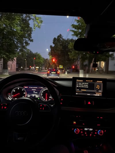 The image shows the interior of a car, looking out through the windshield at a city street at dusk. The dashboard instruments are illuminated, displaying speed, temperature, and other vehicle information. The car's infotainment screen shows a music player interface. Outside, streetlights and traffic lights are visible, along with trees lining the street and buildings in the background. A dark-colored sedan is in front of the car, and other vehicles are visible further down the street. The sky is a deep blue, indicating the transition from day to night. There are no people visible in the image. The scene appears to be a typical urban environment in Orhei, Moldova.
