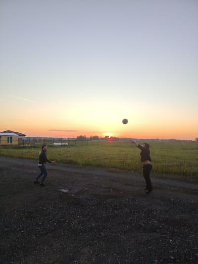 The image depicts two individuals playing volleyball in an open field at dusk, with the sun setting in the background. The sky is a gradient of soft blues and oranges, indicative of a clear evening. The ground is a gravel path, with a green grassy field adjacent to it. In the distance, a small, light-colored house with a dark roof can be seen, along with a wooden fence and some scattered structures.
One person, on the left side of the frame, is a male wearing a black leather jacket and blue jeans. He is in motion, looking up at the ball. The second person, on the right, is also a male wearing dark clothing. He is in mid-air, jumping to hit the ball, which is positioned above his head and slightly to the left.
The scene appears to be a rural or suburban area, likely in Rastunovo, Russia, given the context. The overall mood is relaxed and active, suggesting a casual recreational activity between friends or family. The lighting conditions, with the sun low on the horizon, create a warm and tranquil atmosphere. There is no visible text in the image.