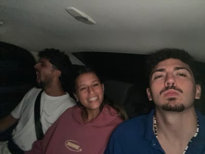 The scene depicts three individuals inside a vehicle at night in Arlington, United States. The vehicle's interior features a light-colored headliner with an oval-shaped overhead light fixture. On the far left, a male with dark, curly hair, wearing a white t-shirt, is captured with his mouth wide open in an apparent shout, facing leftward. A white seatbelt strap is visible across his chest. In the central foreground, a female with long brown hair, wearing a maroon hoodie with "BROWNIE" and a star logo on the front, smiles broadly and looks directly towards the camera. On the right, a male with dark, curly hair, a mustache, and a goatee, dressed in a blue collared shirt and a multi-colored beaded necklace, maintains a direct gaze towards the camera with a neutral expression. The background outside the vehicle is dark.