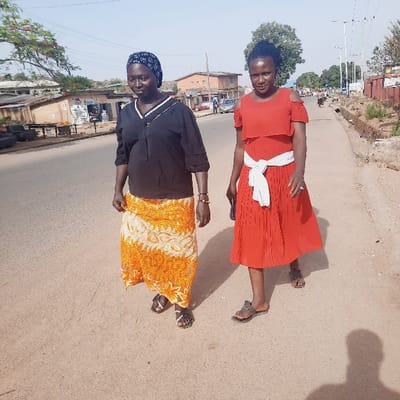 Two women walk down a paved road in the city of Kufang, Nigeria. The woman on the left wears a black blouse and a long yellow and white skirt. She wears sandals. The woman on the right wears a red sleeveless dress with a white sash and sandals. They are walking in the same direction, and the woman on the right appears to be carrying a purse. The road has a sandy surface, and there are buildings on both sides. The sky is blue and clear, and the sun is shining. There is vegetation on the sides of the road.