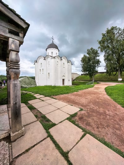 This is a full-shot, eye-level, outdoor, panoramic photograph of the Staraya Ladoga Fortress in Staraya Ladoga, Russia. The sky is overcast and cloudy, with a gray and muted tone. The foreground is dominated by a weathered wooden structure on the left, with rough-hewn posts and a roofline visible. A pathway made of large, square concrete slabs extends from the wooden structure into the scene, with patches of grass growing between the slabs.
To the right of the pathway is a reddish-brown dirt path that curves into the distance. In the middle ground, a white stone church with a dark dome and a cross on top stands prominently. The church has arched windows and decorative architectural details. Adjacent to the church, on the left side of the image, a large green military-style tent is visible. Beyond the church, the fortress walls and grassy slopes are visible, along with several trees with green foliage. A white railing and steps are also present on the right side of the fortress grounds, leading up a grassy incline. Two people are standing near the green tent on the far left of the image, appearing to observe the scene. The overall atmosphere is somber and somewhat damp, typical of a cloudy day.
