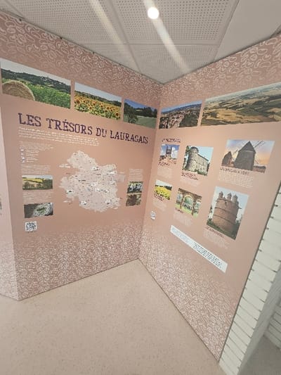 This image displays an interior corner of a room, functioning as an interpretive exhibit, located within a public or informational building in Avignonet-Lauragais, France. The walls are covered with light pinkish-beige panels patterned with numerous small, white, bean-shaped outlines.
The left wall features the prominent title "LES TRÉSORS DU LAURAGAIS" (The Treasures of Lauragais) in dark blue lettering. Above this title, five horizontal rectangular photographs depict various regional landscapes, including a hay bale in a field, a sunflower field, green plains, a hilltop village, and an aerial view of cultivated land. Below the title, a regional map of Lauragais is centrally positioned, flanked by blocks of descriptive text in French and two additional photographs at the bottom showing a bridge structure and a body of water. Two QR codes are present in the lower-left section.
The right wall continues the exhibit theme with multiple rectangular photographs showcasing architectural subjects, specifically historical buildings resembling castles or fortified structures, and at least two windmills. Each image is accompanied by explanatory text in French.
The floor is a light beige color, appearing clean and unadorned. The ceiling consists of a white suspended grid system with perforated acoustic tiles, and a single bright, circular recessed light fixture illuminates the corner from above. The space is unoccupied; no individuals or movable objects are present.