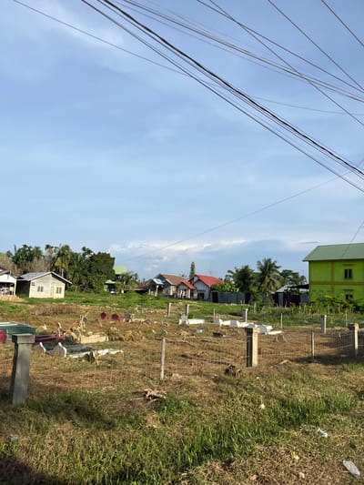 This is an eye-level, outdoor, medium shot of a cemetery in Landuh, Indonesia. The cemetery is in the foreground, with tombstones scattered across dry, patchy grass. A wire fence encloses part of the cemetery, with concrete posts. In the midground, there are several houses with red and green roofs, as well as lush green trees. A bright green, two-story building is prominent on the right side of the image. The background shows a clear blue sky with some scattered white clouds. Utility wires are visible in the upper portion of the image, stretching across the sky. There are no people or animals visible in the scene. The overall impression is one of a rural or semi-rural community.