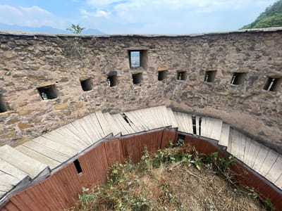 This media file depicts a view from within an ancient stone structure, possibly a fort or castle, looking down at a winding wooden staircase. The stone walls, constructed from rough-hewn rocks of varying sizes and colors, are interspersed with numerous small, rectangular openings or embrasures, suggesting a defensive purpose. These openings offer glimpses of the landscape beyond, hinting at a mountainous or hilly terrain and a distant settlement.

The central focus of the foreground is the descending wooden staircase, which has a curved design and is made of light-colored planks. The staircase is bordered by a dark reddish-brown wooden railing, adding a contrasting color and texture. Overgrown dry grass and green vegetation are present around the base of the staircase and in the interior of the structure, indicating a natural environment encroaching on the man-made elements.

The sky above is partly cloudy, with patches of blue sky visible. The lighting suggests it is daytime, likely late morning or early afternoon. The overall impression is one of historical significance and rustic beauty. There is no visible text in the image. The setting appears to be a historical site in Srinagar, India, given the location context provided. The image captures a sense of age and timelessness, with the sturdy stone structure contrasting with the wooden additions that likely serve as modern access.