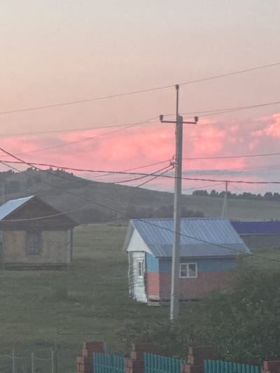 A rural scene in Aumyshevo, Russia, is depicted at dusk. The sky is filled with soft pink and white clouds, suggesting the setting sun. Power lines crisscross the frame, supported by a utility pole. In the midground, two small houses are visible. The house on the left appears to be made of wood with a dark roof and a single window. The house on the right has a blue metal roof and a facade painted in horizontal bands of white, red, and blue. A small white window is visible on this house. The ground is covered in green grass. In the foreground, a section of a brick wall and a green metal fence are present. The background features a rolling hill covered in trees. The overall atmosphere is serene and peaceful.