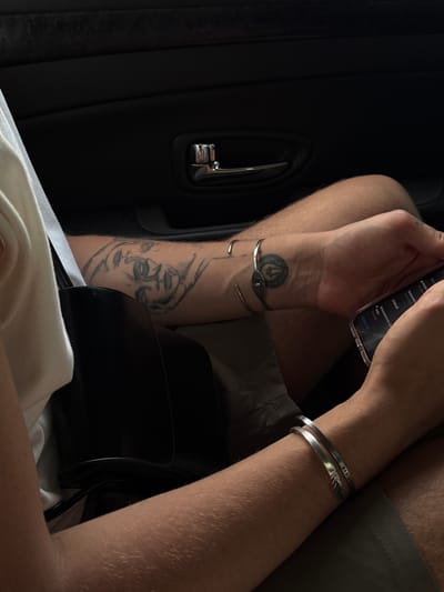 This image depicts a person's arms and upper body, seated in what appears to be the back seat of a car. The background is the dark interior of the car, with a visible door handle and textured surface. The person's left arm has a tattoo of a face. They are wearing two silver bracelets on their right wrist, one of which has an emblem and the other is a simple band with an inscription. The person is holding a smartphone in their right hand. The overall lighting suggests an indoor or shaded environment. There is no indication of the city of Marneuli, Georgia within the image.