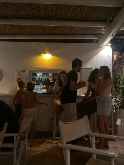 The image depicts an evening scene within a bar or restaurant in Φάρος, Greece, characterized by white-washed walls and a thatched ceiling with white wooden beams. Nine individuals are visible. At the center, a male bartender with a beard, wearing a dark shirt, is positioned behind a well-lit bar counter, actively mixing a drink. Behind him, shelves are stocked with bottles and glassware. To the right of the bartender, a female is seated at the bar, observing a mobile phone. Adjacent to her, a standing male holds a glass and a stirrer, facing two seated females. The female on the left has dark hair, while the female on the right, wearing a light pink top and a light denim skirt, has blonde hair; both are engaged in conversation with the standing male. In the left foreground, two shirtless young boys are standing near the bar area; one, closer to the viewer, wears patterned shorts and faces away, partially obscured by a white chair. The flooring is light-colored and appears to be stone or tile. White-painted wooden furniture, including bar stools and chairs, is present. Green foliage is visible in a planter on the right side of the frame.