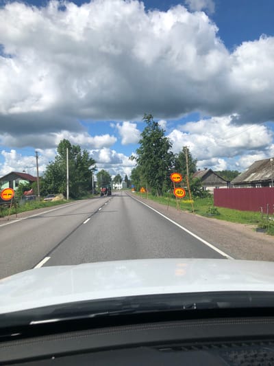 The image captures a view from a vehicle driving on a paved road in Krasnyi Bor, Russia. The road is bordered by a white line on the right and a grassy verge with trees and houses on the left. Several traffic signs are visible along the roadside, including a circular sign with "40" in black on a white background within a red circle, indicating a speed limit of 40. Another sign shows a car with a diagonal red bar through it, possibly indicating a prohibition on certain vehicles.
The scene appears to be set in the daytime, with a bright blue sky interspersed with large, fluffy white and grey clouds, suggesting fair weather with some cloud cover. The landscape is rural, with houses, trees, and a red barn visible. A tractor is also seen on the road ahead. The overall impression is of a sunny day in a countryside setting. The perspective suggests the photo was taken from the passenger seat or dashboard of a moving vehicle, as evidenced by the reflection in the windshield on the right side of the frame.