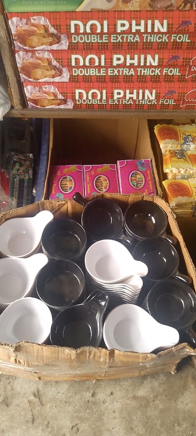 This image depicts a detailed view of merchandise within a retail or market setting in Okere-Urhobo, Nigeria.

The foreground features a rough, grey concrete floor, upon which rests a large, light brown cardboard box. This box is open and partially filled with numerous small, shallow bowls, approximately half glossy black and half matte white, all featuring an integrated handle-like protrusion. These bowls are stacked in multiple columns within the box.

Above and behind this box, a display area is visible. On a higher shelf, at least three rolls of "DOLPHIN DOUBLE EXTRA THICK FOIL" are horizontally stacked. The foil packaging is red with a tartan pattern, displaying images of cooked chicken in foil and a small purple dolphin logo. Below this, on a lower shelf or within another open box, are several pink boxes labeled "100 Cocktail Parasols," featuring colorful illustrations of fruit slices and umbrellas. To the far right, stacks of yellow packaging with "TAKE HOME" text and cartoon characters resembling Tom and Jerry are partially visible, likely representing party napkins or small bags. The left side of the background shows indistinct, dark, plastic-wrapped items and a blue object. No individuals are present in the scene.
