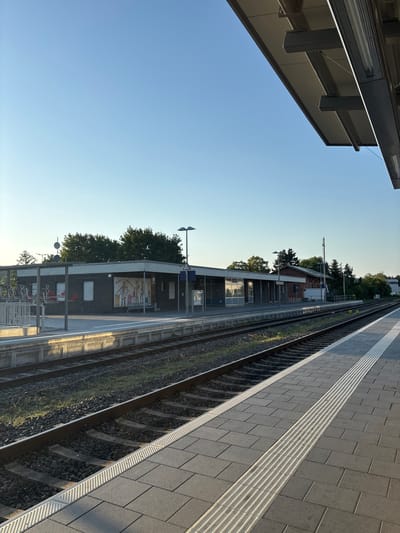 This is a landscape photograph taken at a train station in Elsenfeld, Germany. The image is oriented vertically. On the left side of the frame, a modern building with dark gray cladding and large windows is partially visible. The building casts a shadow on the clear blue sky.
The right side of the image shows a train platform with textured gray tiles. A tactile paving strip runs along the edge of the platform next to the train tracks. The tracks themselves are filled with ballast and bordered by green grass.
In the background, beyond the tracks, is a small brick building with a pitched roof, likely a station house. Trees line the background, suggesting a suburban or semi-rural setting. A street light is visible on a pole in front of the brick building. The sun is low in the sky, casting long shadows and creating a warm, golden light that reflects on the windows of the station building. This indicates that the photograph was taken either in the early morning or late afternoon.
Some visible text on the brick building reads "Herzlich" (meaning "cordially" or "warmly" in German), possibly part of a welcome sign. Other text is illegible. There are no people visible in the image, and the scene appears quiet and serene.