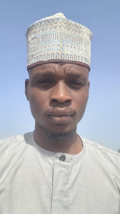 A frontal close-up portrait displays an adult male outdoors in Badiko, Nigeria, under a clear, light blue sky. The individual has a dark skin tone, a short dark beard, and a mustache, with a neutral expression. He wears a light-colored traditional cap featuring intricate blue and beige geometric embroidery and a dark red band around the base. He is dressed in a light gray long-sleeved garment with a band collar and a partial placket, secured by one visible dark button. Bright, direct natural light illuminates the subject, causing a sheen on his forehead and face. No other individuals or specific environmental features are discernible.