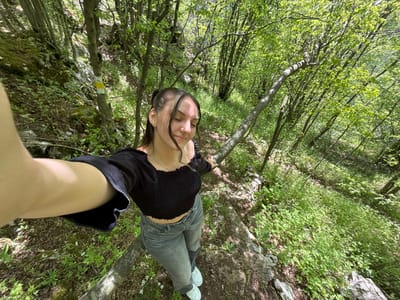 A young woman is lying on a rocky outcrop in a forest. She is wearing jeans, a black crop top, and white sneakers. Her eyes are closed. The forest consists of tall trees with green leaves, and the ground is covered with vegetation. There is a tree with a yellow and white trail marker on it. The location is Lipnitsa, Bulgaria.