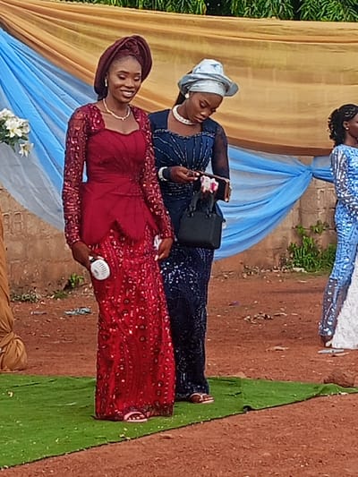 The scene, set outdoors in Afaka, Nigeria, depicts three young adult women. The woman on the left wears a maroon long-sleeved, floor-length gown adorned with sequins, a matching maroon gele, a gold-colored beaded necklace, and white earrings. She exhibits a slight smile and holds a white handheld fan in her left hand. Adjacent to her, a second woman is dressed in a dark blue sequined long-sleeved gown, a light blue hat-style gele, a white choker necklace, and white earrings. She is observed looking down at a brown wallet or clutch held in her right hand, with a small black handbag hanging from her left. Partially visible on the far right, a third woman wears a light blue sequined long-sleeved dress. The background features draped decorative fabrics in gold and light blue, partially obscuring green foliage and a light brown structure. The ground consists of reddish-brown dirt, over which a strip of artificial green turf is laid. White floral arrangements are visible to the far left.